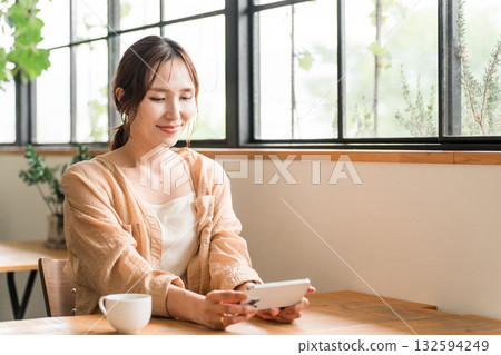 Young Asian woman drinking coffee in the living room of a cafe and looking at her smartphone Young Asian woman drinking coffee in the living room of a cafe and looking at her smartphone 132594249