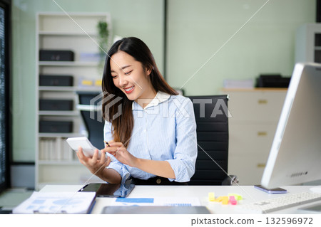 Happy Asian woman calculates business finances in a bright office. Modern accounting, planning, and real people concept 132596972