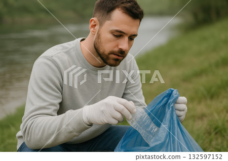 Man wearing gloves carefully cleaning riverbank by picking up plastic waste with a blue trash bag on a cloudy outdoor day, AI Generative 132597152