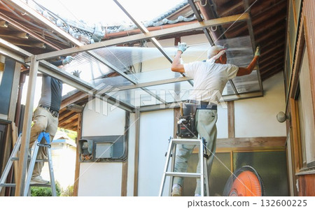 Two carpenters lifting a polycarbonate corrugated sheet to install it on the roof Two carpenters lifting a polycarbonate corrugated sheet to install it on the roof 132600225