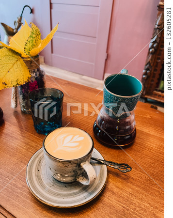 Latte art coffee is served on wooden table next to empty decorated oriental coffee pot and blue glass, with autumn leaves in background giving cozy atmosphere. Latte art, oriental coffee, v60, cafe 132605281