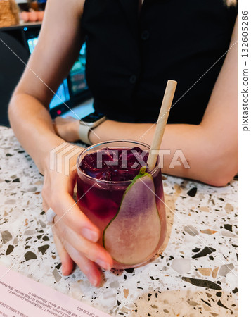 Woman is holding colorful cocktail with ice cubes, pear slice, and paper straw at modern bar, enjoying refreshing drink in stylish setting. Vibrant cocktail with pear garnish, ice, paper straw Woman is holding colorful cocktail with ice cubes, pear slice, and paper straw at modern bar, enjoying refreshing drink in stylish setting. Vibrant cocktail with pear garnish, ice, paper straw 132605286