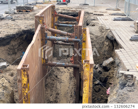 Busy urban construction site with a deep excavated trench, securely supported by a heavy-duty metal shoring system, for essential underground utility line installation 132606274