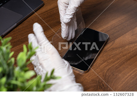Gloved hands meticulously applying a clear screen protector onto a sleek smartphone on a wooden desk, ensuring precise gadget protection 132607328