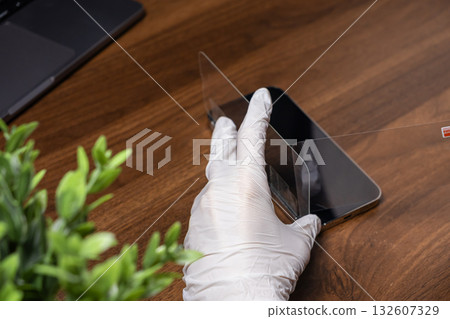 Gloved hand carefully applies clear protective FRONT screen film to a smartphone display on wooden desk, enhancing device durability and safeguarding privacy 132607329