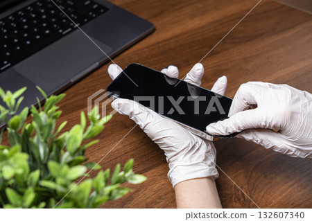 Gloved hands diligently sanitizing smartphone screen with a soft cloth on a wooden desk, promoting hygiene for safe daily device usage Gloved hands diligently sanitizing smartphone screen with a soft cloth on a wooden desk, promoting hygiene for safe daily device usage 132607340