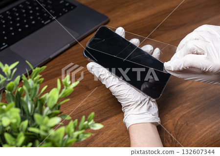 Gloved hands carefully removing a cracked tempered glass screen protector from a modern smartphone, illustrating professional mobile repair services Gloved hands carefully removing a cracked tempered glass screen protector from a modern smartphone, illustrating professional mobile repair services 132607344