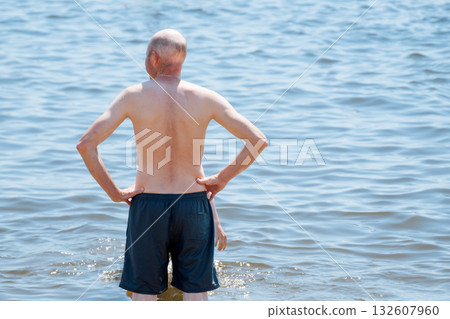 A bald man stands in swimming trunks in the sea and sunbathes, looking away A bald man stands in swimming trunks in the sea and sunbathes, looking away 132607960