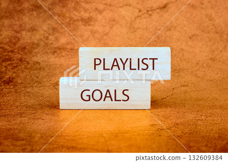 Two wooden blocks are stacked on a textured surface. The top block says PLAYLIST and the bottom block says GOALS. They represent musical aspirations on an orange background Two wooden blocks are stacked on a textured surface. The top block says PLAYLIST and the bottom block says GOALS. They represent musical aspirations on an orange background 132609384