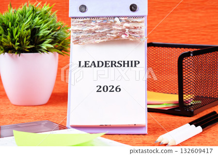 A desk calendar displays LEADERSHIP and 2026. Various desk supplies, a potted plant, and a black wire mesh organizer are arranged on an orange surface A desk calendar displays LEADERSHIP and 2026. Various desk supplies, a potted plant, and a black wire mesh organizer are arranged on an orange surface 132609417