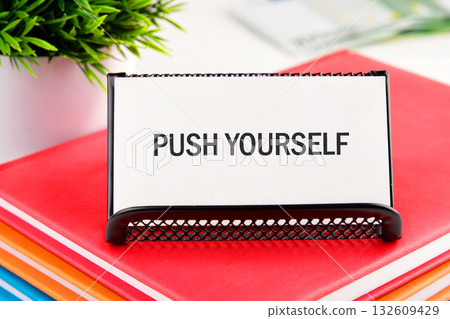A rectangular white sign with the words PUSH YOURSELF rests on colorful books. A small green potted plant sits nearby, adding a touch of nature to the inspiring scene A rectangular white sign with the words PUSH YOURSELF rests on colorful books. A small green potted plant sits nearby, adding a touch of nature to the inspiring scene 132609429