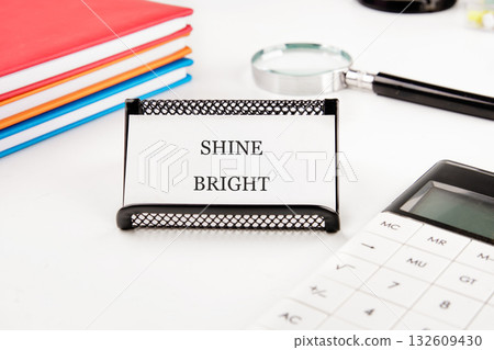 On a white desk, a black holder displays a card with the words SHINE BRIGHT. Surrounding it are colorful notebooks, a magnifying glass, and a white calculator in a bright, clean workspace On a white desk, a black holder displays a card with the words SHINE BRIGHT. Surrounding it are colorful notebooks, a magnifying glass, and a white calculator in a bright, clean workspace 132609430