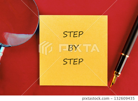 A yellow sticky note with the words STEP BY STEP is placed on a red background next to a pen and magnifying glass, suggesting a methodical approach A yellow sticky note with the words STEP BY STEP is placed on a red background next to a pen and magnifying glass, suggesting a methodical approach 132609435
