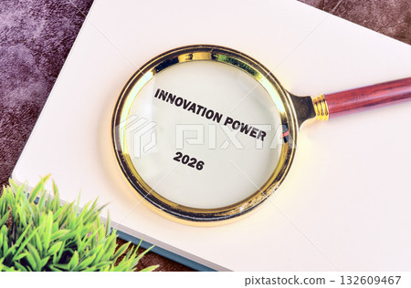 A magnifying glass rests on a notepad revealing the words INNOVATION POWER 2026. A small artificial green plant is near the notepad against a dark background 132609467
