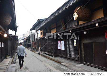 Old townscape of Takayama City, Gifu Prefecture, Japan. A sake shop decorated with cedar balls and a man taking a walk. Morning scene during the Takayama Festival in spring. Old townscape of Takayama City, Gifu Prefecture, Japan. A sake shop decorated with cedar balls and a man taking a walk. Morning scene during the Takayama Festival in spring. 132609540