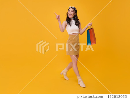 Happy young Asian woman shopper walking carrying shopping bags pointing finger up isolated on yellow copy space background. Happy young Asian woman shopper walking carrying shopping bags pointing finger up isolated on yellow copy space background. 132612053