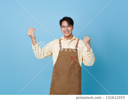 smiling Asian man in apron gestures happily with both hands exuding confident and successful demeanor isolated on blue background. 132612055