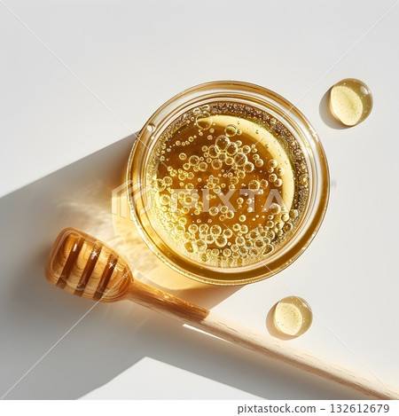 Minimal food flatlay: a round glass jar filled with golden honey covered in tiny bubbles, with a wooden honey dipper and glossy drops scattered on a white surface. Crisp texture and natural color for  132612679