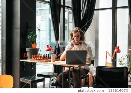 Young man working on laptop in modern cafe while talking on the phone Young man working on laptop in modern cafe while talking on the phone 132614073