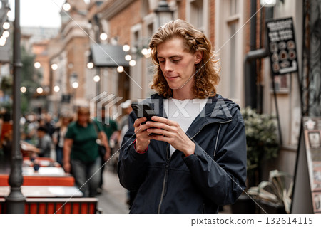 Young man uses smartphone while standing in busy street alley during daytime Young man uses smartphone while standing in busy street alley during daytime 132614115