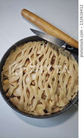 Homemade apple pie with lattice crust ready for baking placed beside a knife and rolling pin. The image conveys the concept of traditional dessert and cozy home cooking 132616412