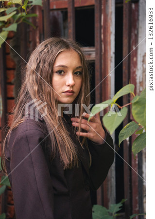 Teen girl with wavy hair stands near rusty metal bars, surrounded by green leaves, in soft natural light Teen girl with wavy hair stands near rusty metal bars, surrounded by green leaves, in soft natural light 132616933