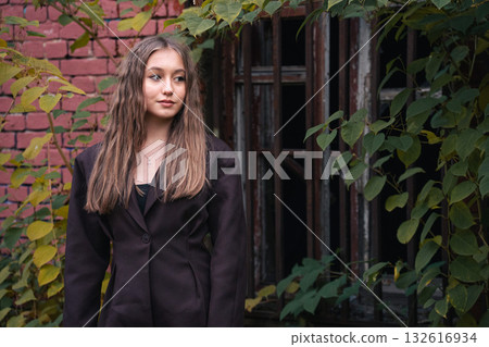 Teen girl with wavy hair stands near rusty metal bars, surrounded by green leaves, in soft natural light Teen girl with wavy hair stands near rusty metal bars, surrounded by green leaves, in soft natural light 132616934