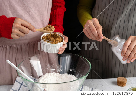 Two people preparing baking ingredients adding brown sugar and salt to flour in a glass bowl. Holiday cookie baking scene in a cozy kitchen 132617870