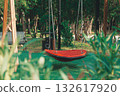 Bright red swing in a lush green park surrounded by trees and benches during a sunny day 132617920
