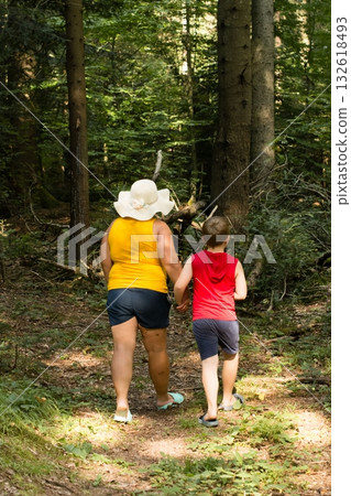 A woman and child stroll through a sun-dappled forest path, enjoying a leisurely walk amongst the trees. 132618493