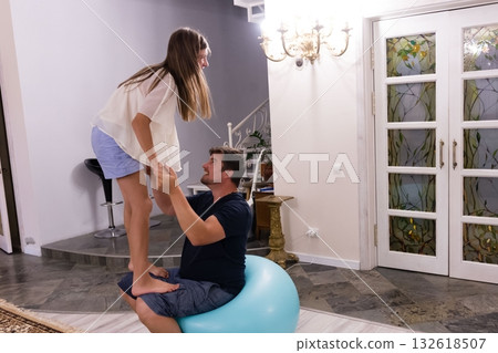 Father and daughter playfully interact, with the girl standing on an exercise ball while her dad assists. 132618507