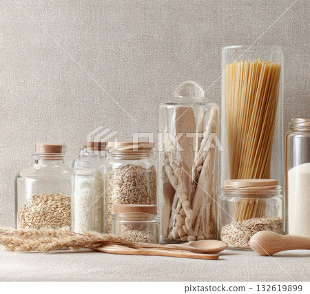 Glass jars with dried food, pasta, rice, oat, wheat, and wooden utensil on beige fabric background create cozy, natural kitchen storage scene with neutral tones and organic textures 132619899