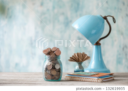 Jar of coins, recycled notebook, blue desk lamp, and paper fan on wooden table, eco friendly finance concept with soft natural light and calm atmosphere 132619935