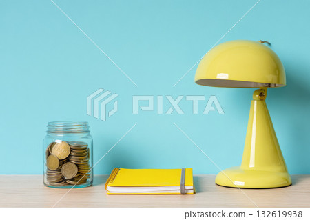 Jar of coins, recycled notebook, and yellow desk lamp on wooden table with blue background, symbolizing eco friendly finance and sustainable workspace with cheerful mood Jar of coins, recycled notebook, and yellow desk lamp on wooden table with blue background, symbolizing eco friendly finance and sustainable workspace with cheerful mood 132619938