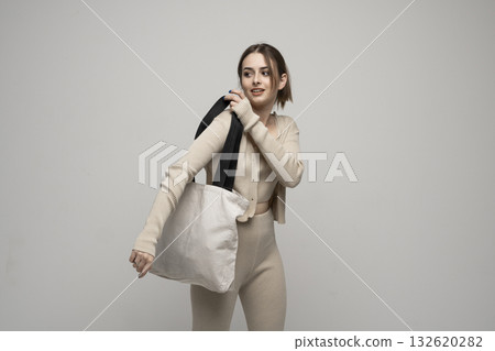Stylish young woman in beige outfit holding eco fabric bag on shoulder in studio 132620282