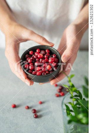 Hand holding bowl of fresh cranberries on a light table with green branch. Hand holding bowl of fresh cranberries on a light table with green branch. 132621302