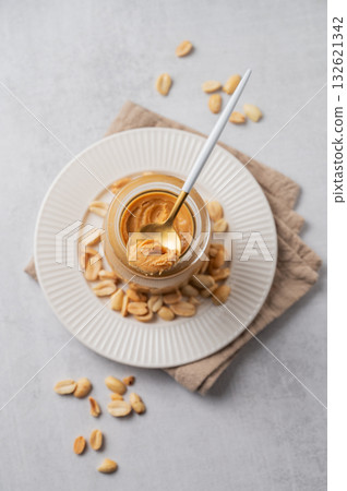 Creamy peanut butter in a jar on a light background with scattered roasted nuts, spoon Creamy peanut butter in a jar on a light background with scattered roasted nuts, spoon 132621342