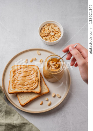 A hand holds a spoon with creamy peanut butter in a jar on a plate with toast and roasted nuts A hand holds a spoon with creamy peanut butter in a jar on a plate with toast and roasted nuts 132621343