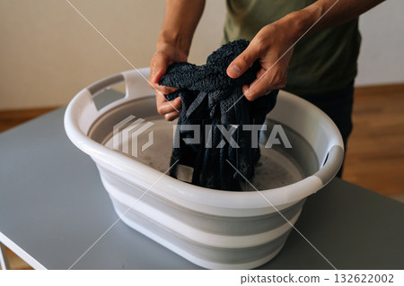 Close-up of unrecognizable man squeezing water from black garment while washing clothes by hand in plastic basin placed on table at home, representing sustainable and traditional laundry method Close-up of unrecognizable man squeezing water from black garment while washing clothes by hand in plastic basin placed on table at home, representing sustainable and traditional laundry method 132622002