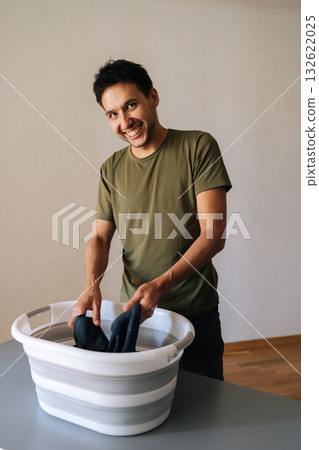Vertical portrait of cheerful young man washing clothes by hand in plastic basin, wringing out water performing domestic task, smiling looking at camera. Concept of hygiene and cleanliness. Vertical portrait of cheerful young man washing clothes by hand in plastic basin, wringing out water performing domestic task, smiling looking at camera. Concept of hygiene and cleanliness. 132622025