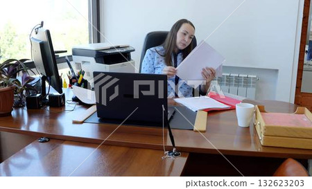 Focused businesswoman reviewing paperwork in a busy office, surrounded by equipment and supplies. Natural light creates a productive atmosphere for efficient work Focused businesswoman reviewing paperwork in a busy office, surrounded by equipment and supplies. Natural light creates a productive atmosphere for efficient work 132623203