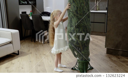 Young girl with golden hair caressing illuminated christmas tree wrapped in protective mesh, standing inside contemporary apartment, embodying festive warmth Young girl with golden hair caressing illuminated christmas tree wrapped in protective mesh, standing inside contemporary apartment, embodying festive warmth 132623224
