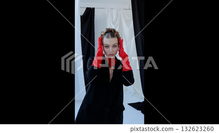 High fashion model posing in studio, wearing crimson gloves and black blazer. Piercing stare against monochrome backdrop, exuding sophisticated elegance and dramatic styling 132623260