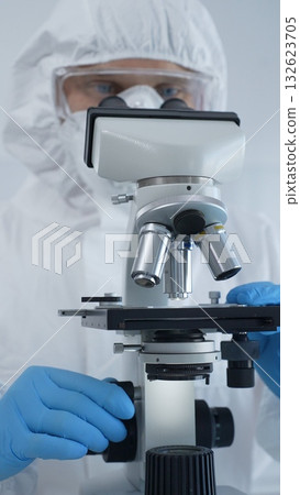 Male virologist in a hazmat suit, mask, and blue gloves, diligently working at a microscope in a laboratory researching viruses. Medicine, science concept 132623705