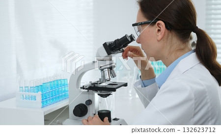 Female scientist examining microscope slide, analyzing chemical sample in sterile laboratory environment, performing precision medical research. Medicine and science concept Female scientist examining microscope slide, analyzing chemical sample in sterile laboratory environment, performing precision medical research. Medicine and science concept 132623712