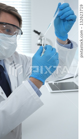 Masked male scientist in lab coat, protective glasses and blue gloves is adding reagent to test tube with microscopes and test tube rack in modern laboratory. Medicine, healthcare and science concept 132623719