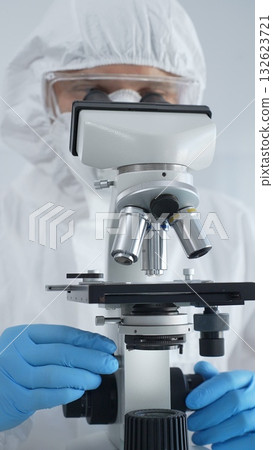 Male virologist in a hazmat suit, mask, and blue gloves, diligently working at a microscope in a laboratory researching viruses. Medicine, science concept Male virologist in a hazmat suit, mask, and blue gloves, diligently working at a microscope in a laboratory researching viruses. Medicine, science concept 132623721