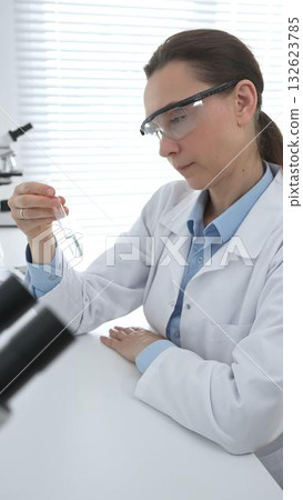 Female researcher examining clear liquid in test tube, sitting near microscopes and lab equipment within sterile scientific workspace. Medicine, healthcare and science concept Female researcher examining clear liquid in test tube, sitting near microscopes and lab equipment within sterile scientific workspace. Medicine, healthcare and science concept 132623785