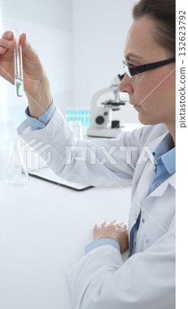 Female researcher examining clear liquid in test tube, sitting near microscopes and lab equipment within sterile scientific workspace. Medicine, healthcare and science concept 132623792