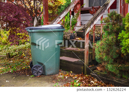 Large green garbage bin on wheels standing near the front steps of an old wooden porch surrounded by autumn foliage. Large green garbage bin on wheels standing near the front steps of an old wooden porch surrounded by autumn foliage. 132626224